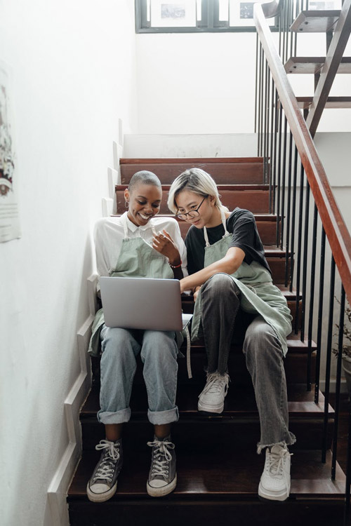 Two black women sit on a staircase looking at a computer.