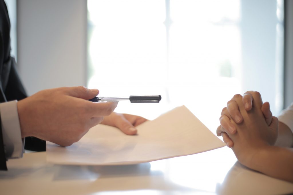 Person handing pen across table to person with hands folded